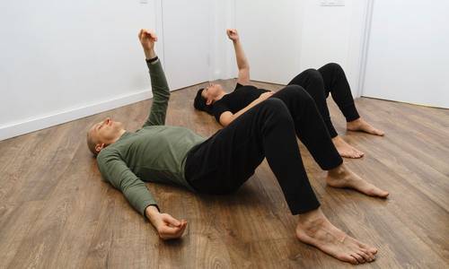 Zwei Personen liegen auf dem Holzboden und machen Gymnastik