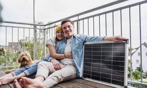 Ein nunges Paar mit Hund sitzt auf dem Boden ihres Balkons, daneben ein Solarmodul