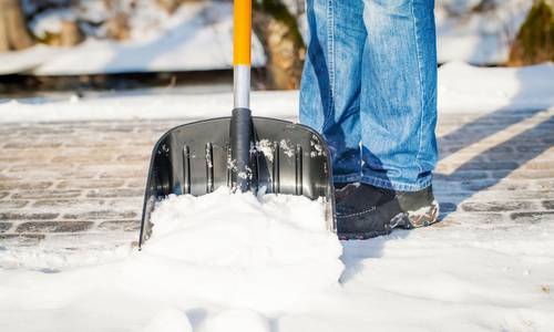 Ein Ausschnitt einer Schneeschaufel, mit der Schnee auf einem Gehweg geräumt wird