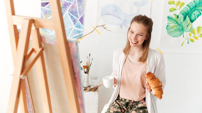 Frau sitzt lachend vor Staffelei und hält eine Tasse Kaffee in der Hand